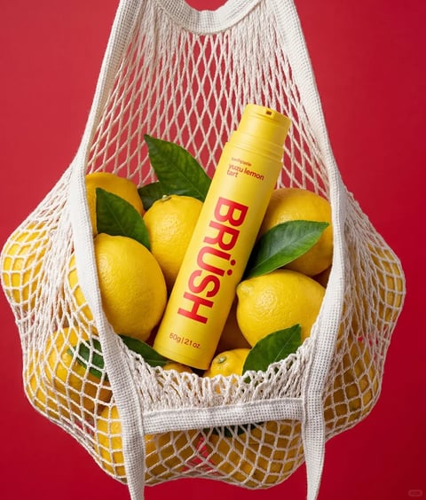 The image is a high-contrast, vibrant product photograph featuring a yellow tube of toothpaste nestled among fresh lemons inside a white mesh produce bag, all set against a solid, saturated red background. The composition is centered, with the bag and its contents filling the frame, creating a bold, graphic arrangement. The toothpaste tube is the focal point, positioned diagonally among the lemons, which are bright yellow with glossy, textured rinds and some attached dark green leaves. Visible text on the tube includes the brand name 'BRUSH' in large, bold, red sans-serif letters running vertically, with smaller text above reading 'toothpaste' and 'yuzu lemon tart,' and weight information '50g | 2.1 oz.' at the bottom. The lighting is bright and directional, coming from the upper left, casting soft shadows that define the round forms of the lemons and the cylindrical tube, enhancing their three-dimensionality. The visual aesthetic is clean, modern, and photorealistic, with a strong emphasis on color blocking between the red background, yellow objects, and white bag. The color language is dominated by a high-saturation primary palette of red and yellow, with green accents from the leaves, creating a cheerful, energetic mood. Textures are rendered with clarity: the knitted, porous weave of the cotton bag, the dimpled, waxy skin of the lemons, and the smooth, matte plastic of the tube. The composition uses the organic, rounded shapes of the fruit to contrast with the geometric, cylindrical form of the tube, all contained within the flexible, net-like structure of the bag, which adds a layer of visual texture and depth.
