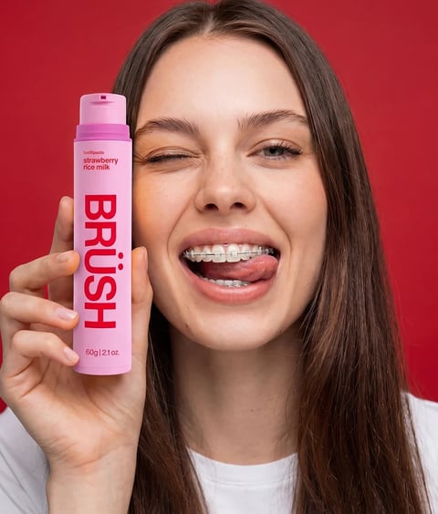 This is a photorealistic, studio-style portrait of a young woman with long, straight brown hair, set against a solid, vibrant red background. The composition is a tight close-up, focusing on her face and the hand holding a product. She has a playful, energetic expression, winking her right eye while sticking her tongue out to lick the corner of her mouth, revealing metal braces on her upper and lower teeth. Her skin is clear with a natural, dewy finish, and her eyebrows are well-defined. In her left hand, held vertically beside her cheek, is a matte pink cylindrical tube of toothpaste. The product's text is clearly visible: the brand name 'BRUSH' is printed vertically in large, bold, red sans-serif letters, with a stylized umlaut over the 'U'. Above this, in smaller red text, are the words 'toothpaste' and 'strawberry rice milk'. At the bottom, the net weight '60g | 2.1 oz.' is printed in small red text. The lighting is bright, even, and frontal, creating soft, minimal shadows and a high-key, commercial feel that emphasizes the product and the subject's vibrant expression. The color palette is dominated by the bold red background, the soft pink of the product tube, and the natural tones of the subject's skin and hair, creating a high-contrast, saturated, and youthful aesthetic. The texture of the matte tube contrasts with the slight sheen of the subject's lips and the metallic glint of her braces.