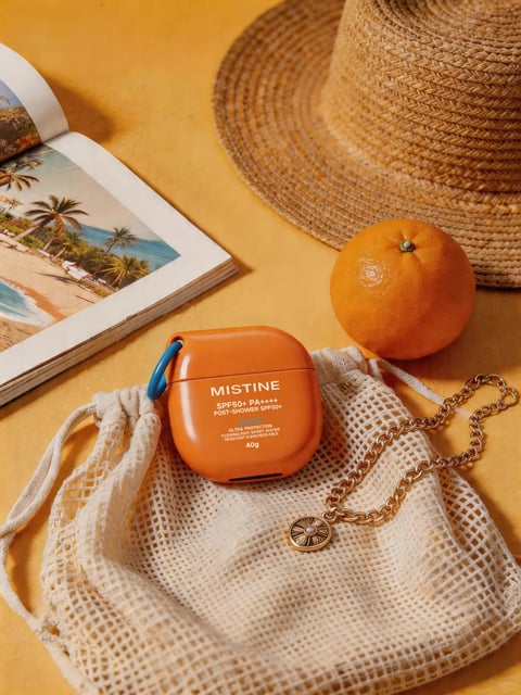 This is a photorealistic, warm-toned still life composition arranged on a smooth, matte mustard-yellow surface, evoking a vibrant, sun-drenched summer mood. The central subject is a glossy, rounded rectangular orange container with a small blue carabiner clip, positioned atop a cream-colored, open-weave mesh drawstring bag. To the left, an open magazine displays a tropical beach scene with palm trees and turquoise water. In the upper right, a textured straw sun hat and a whole, glossy orange fruit add organic shapes and reinforce the seasonal theme. A delicate gold chain necklace with a circular sunburst pendant drapes across the mesh bag. The lighting is bright, directional, and slightly hard, coming from the upper left, casting distinct, warm shadows that enhance the three-dimensionality of the objects. The color palette is dominated by saturated oranges, warm yellows, and creamy neutrals, with accents of blue from the carabiner and the magazine image. Visible text on the orange container reads 'MISTINE', 'SPF50+ PA++++', 'POST-SHOWER SPF50+', 'BODY', 'ULTRA PROTECTION', 'CLEAR&LIGHT, SWEAT & WATER RESISTANT SUNSCREEN MILK', and '40g', all in white sans-serif fonts, clearly identifying the product and its specifications. The composition is tightly framed with a shallow depth of field, keeping the foreground objects sharp while the background hat softens slightly, creating an intimate, focused arrangement with a balance of geometric (container, magazine) and organic (fruit, hat, bag) shapes.