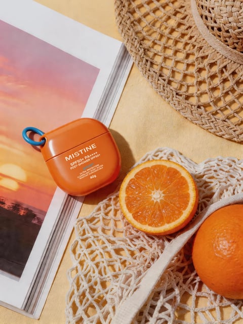 The image is a photorealistic, top-down flat lay composition with a warm, summery aesthetic. The main subject is an orange, rectangular sunscreen container with a blue carabiner clip, positioned slightly left of center on a magazine page featuring a sunset photograph. To the right, a halved orange rests on a beige, knitted mesh bag, with a whole orange partially visible in the bottom right corner. A woven straw hat occupies the upper right background. The color palette is dominated by vibrant oranges, warm yellows, and earthy beiges, with a pop of blue from the clip, creating a high-saturation, high-contrast look with a warm color temperature. The lighting is bright and directional, coming from the upper left, casting soft, defined shadows that enhance the three-dimensionality of the objects. Text is clearly visible on the sunscreen container, reading 'MISTINE', 'SPF50+ PA++++', 'POST-SHOWER SPF50+', 'ULTRA PROTECTION CLEAR & LIGHT SPRAY WAX', 'WATER RESISTANT SUNSCREEN MILK', and '40g' in white sans-serif font, serving as product branding and information. The composition uses overlapping elements and a shallow depth of field, with the sunscreen and orange slice in sharp focus while the background hat is slightly softer. The visual aesthetic combines organic shapes (oranges, hat weave) with the geometric form of the container, creating a tactile, natural, and refreshing mood evocative of summer and outdoor leisure.