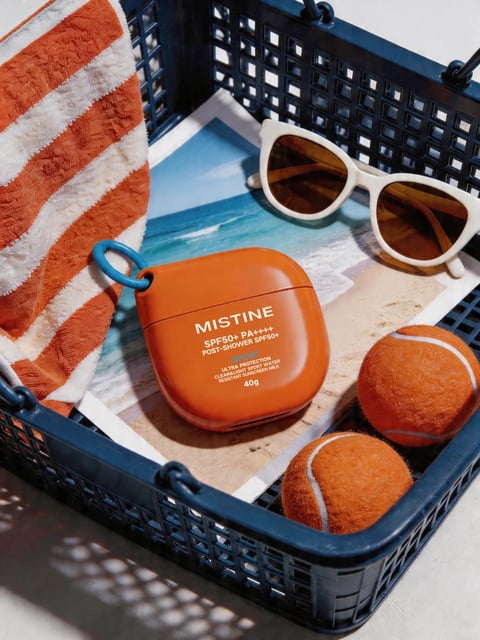 The image is a photorealistic, high-contrast still life composition arranged within a dark blue plastic crate, evoking a vibrant summer or beach-day mood. The main subject is an orange, square-shaped MISTINE sunscreen container with rounded edges and a blue carabiner clip, placed centrally on a photographic print of a sunny beach scene. Surrounding it are two fuzzy orange tennis balls, a pair of white-framed sunglasses with amber lenses, and a folded orange-and-white striped towel draped over the crate's edge. The visible text on the sunscreen container is in English, reading 'MISTINE SPF50+ PA++++ POST-SHOWER SPF50+ DRY ULTRA PROTECTION CLEAR&LIGHT SPORT WATER RESISTANT SUNSCREEN MILK 40g' in white sans-serif font, clearly identifying the product and its features. The lighting is bright and directional, coming from the upper left, casting sharp, defined shadows that enhance the three-dimensionality of the objects and create a warm, sun-drenched atmosphere. The color palette is dominated by saturated oranges and warm tones, contrasted with the deep blue of the crate and the cool blues of the beach photograph, creating a high-energy, playful, and tactile visual experience with a focus on summer leisure and protection.