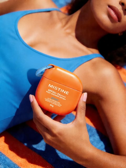 The image presents a close-up, lifestyle-style photograph featuring a person's upper torso and hand holding an orange sunscreen container. The composition is framed from a slightly high angle, focusing on the product held against the person's chest, with their face partially visible in the upper right corner, cropped at the nose. The person wears a vibrant blue athletic tank top, and their skin has a warm, sun-kissed tone. The background consists of a blue and orange striped towel or fabric, creating a cohesive color scheme. The visual aesthetic is photorealistic with a clean, commercial quality, emphasizing a bright, summery mood. The color language is dominated by a high-saturation contrast between the vivid cerulean blue of the top and towel stripes and the bright, warm orange of the sunscreen container and towel stripes, with the person's skin adding warm peach and tan tones. The lighting is bright and directional, appearing to be natural sunlight coming from the upper left, casting soft shadows that define the contours of the hand, collarbone, and product, conveying a warm, outdoor atmosphere. Texture and material feel are rendered with clarity: the smooth, matte plastic of the orange sunscreen bottle, the soft, slightly textured fabric of the blue tank top, the plush, looped texture of the towel, and the natural, smooth texture of the skin. Compositionally, the image uses a shallow depth of field, keeping the product and hand in sharp focus while the background and parts of the person's face and body are softly blurred, directing attention to the held object. Shape language combines the organic curves of the person's body and hand with the geometric, rounded-square shape of the sunscreen container and its attached blue carabiner clip. The visible text on the orange container is printed in white sans-serif font and reads 'MISTINE' at the top, followed by 'SPF50+ PA++++', 'POST-SHOWER SPF50+', 'SPORT', 'ULTRA PROTECTION', 'CLEAR&LIGHT SPORT WATER', 'RESISTANT SUNSCREEN MILK', and '40g' at the bottom; this text is central to the product's branding and informational role. The overall mood is energetic, fresh, and summery, evoking themes of outdoor activity, sun protection, and vibrant leisure.