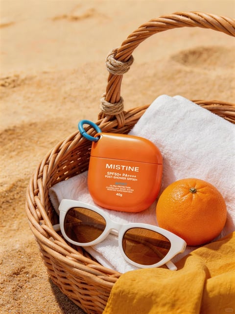 This is a photorealistic, high-resolution lifestyle product photograph composed in a close-up, slightly high-angle shot, focusing on a curated summer beach essentials arrangement inside a woven wicker basket set against a sandy background. The main subject is an orange, rounded-square sunscreen container from the brand MISTINE, positioned centrally within the basket alongside a fresh orange, a pair of white-framed sunglasses with brown lenses, and a folded white towel, with a mustard-yellow fabric draped over the basket's edge. The composition uses a shallow depth of field, keeping the basket's contents in sharp focus while the sandy background softens into a warm, textured blur. The visible text on the orange container is printed in clean, white, sans-serif English typography, reading "MISTINE" prominently at the top, followed by "SPF50+ PA++++", "POST-SHOWER SPF50+", and smaller descriptive text below including "ULTRA PROTECTION", "CLEAR & LIGHT SPORT WATER RESISTANT SUNSCREEN MILK", and "40g", clearly identifying the product and its specifications. The visual aesthetic is clean, modern, and aspirational, with a rendering style that emphasizes realistic textures and materials. The color language is dominated by a warm, saturated palette of vibrant orange, sandy beige, crisp white, and mustard yellow, creating a cohesive, sun-drenched feel with high contrast between the bright objects and their soft shadows. The lighting approach utilizes strong, directional natural sunlight coming from the upper left, casting distinct, hard-edged shadows that define the forms and textures, conveying a bright, midday outdoor mood. Texture and material feel are highly detailed, showcasing the rough, granular quality of the sand, the intricate woven pattern of the wicker basket, the soft terry cloth of the towel, the smooth matte plastic of the sunscreen container, and the dimpled peel of the orange. The composition follows a natural, slightly asymmetrical arrangement within the basket's circular frame, creating a sense of casual abundance and depth through overlapping objects. Shape language combines the organic curves of the basket handle, orange, and sunglasses with the geometric, rounded-square form of the sunscreen container. The overall mood and atmosphere are cheerful, refreshing, and evocative of a relaxing, sunny day at the beach, with a high energy level conveyed through the vibrant colors and bright lighting.