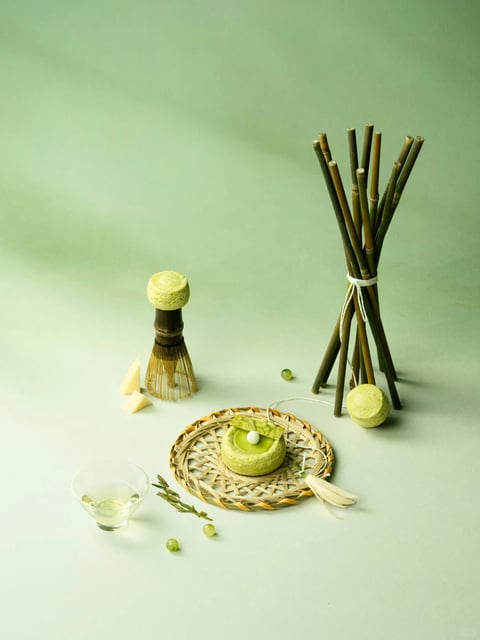The image presents a minimalist, photorealistic still life composition centered on a light sage-green surface and backdrop, featuring a curated arrangement of objects that evoke a natural, artisanal aesthetic. The main subject is a collection of items including a small, round, pale green cake or confection with a glossy top and a white spherical garnish, placed on a woven bamboo mat; a traditional bamboo tea whisk (chasen) with a textured, pale yellow top; a small clear glass bowl containing a light liquid; a bundle of thin, dark bamboo stalks tied with white string; and scattered small green grapes and pale yellow triangular pieces. There is no visible text or readable words in the image. The visual aesthetic is clean and refined, with a photorealistic rendering technique that emphasizes subtle textures and natural materials. The color language is dominated by a muted, monochromatic palette of sage greens, pale yellows, and natural bamboo browns, with low saturation and soft contrast, creating a calm, earthy temperature. The lighting approach is soft and diffused, appearing to come from the upper left, casting gentle, elongated shadows that enhance the three-dimensional form of the objects without harsh edges. Texture and material feel are highlighted through the tactile rendering of the woven bamboo, the fibrous tea whisk, the smooth glass, and the slightly porous surface of the confection. Composition patterns follow a balanced, asymmetrical arrangement with a central focal point on the cake, using negative space to create a sense of openness and depth. Shape language combines organic forms (grapes, cake) with geometric elements (triangular pieces, cylindrical bamboo stalks), featuring soft edges and delicate line weights. The overall mood and atmosphere convey tranquility, simplicity, and a connection to nature, with a low-energy, contemplative visual weight.