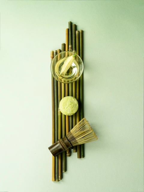This is a minimalist, photorealistic flat-lay composition arranged vertically against a smooth, pale mint-green background. The main subject is a curated set of objects associated with Japanese matcha tea preparation, laid upon a central column of parallel, natural bamboo stalks of varying green and tan hues. From top to bottom, these elements include: a small, clear glass bowl containing a pale, translucent flower bud floating in a clear liquid; a round, textured, pale-green matcha sweet (wagashi); and a traditional bamboo matcha whisk (chasen) with a dark-stained handle and fine, light-colored tines. The composition is strictly vertical and centered, with the bamboo stalks creating a strong linear structure. There is no visible text or readable words in the image. The lighting is soft, diffused, and even, coming from the front or slightly above, casting minimal, soft shadows that give the objects a gentle three-dimensionality without harsh contrast. The color palette is muted and harmonious, dominated by natural earth tones—various shades of bamboo green, tan, and brown—accented by the pale green of the sweet and the clear glass, all set against the cool, desaturated mint backdrop. Textures are rendered with high fidelity: the smooth, reflective glass, the fibrous and segmented surface of the bamboo stalks, the porous, slightly crumbly appearance of the wagashi, and the delicate, hair-like tines of the whisk. The overall mood is serene, orderly, and contemplative, emphasizing natural materials, craftsmanship, and a wabi-sabi aesthetic through its clean lines, organic shapes, and tranquil, low-saturation color scheme.