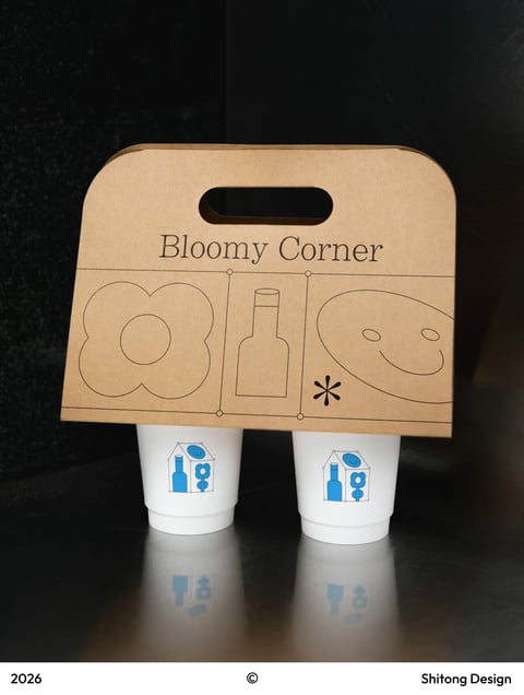 The image features a product packaging design, showcasing a two-cup carrier made of light brown kraft paper, held against a dark, reflective surface that creates a subtle mirror effect. The carrier has a die-cut handle at the top and is divided into three illustrated panels: a stylized flower shape, a simple bottle outline, and a smiling face, all rendered in thin, black line art. Below the carrier, two white paper cups are visible, each printed with a small blue graphic of a house containing a bottle and a flower. The overall composition is centered and symmetrical, with the carrier dominating the upper half and the cups anchoring the lower half, creating a clean, balanced layout. The text 'Bloomy Corner' is printed in a serif font on the upper portion of the carrier, serving as the brand name. The visual aesthetic is minimalist and illustrative, with a flat, graphic style for the line art and a photorealistic rendering of the physical materials. The color language is dominated by the natural, muted brown of the kraft paper, the stark white of the cups, and the deep black background, with a pop of bright blue on the cup graphics. The lighting is soft and directional, coming from the front-left, casting gentle shadows that emphasize the texture of the paper and the three-dimensionality of the carrier and cups. The texture and material feel are highlighted, with the matte, fibrous surface of the kraft paper contrasting with the smooth, slightly glossy finish of the white cups. The shape language combines organic curves in the flower and smiley face with the geometric lines of the bottle and panel divisions, creating a playful yet structured feel. The mood is cheerful, approachable, and modern, evoking a sense of casual, friendly branding for a café or beverage service.