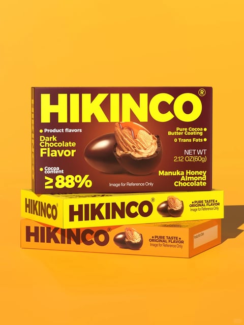 The image presents a stacked arrangement of three rectangular product boxes against a solid, warm yellow-orange background, creating a clean, commercial product shot. The top box, oriented horizontally, is the primary focus, featuring a dark brown base color with bold, bright yellow typography. Its front face displays the brand name 'HIKINCO' in large, sans-serif capital letters at the top. Below this, on the left, text reads 'Product flavors' followed by 'Dark Chocolate Flavor' and 'Cocoa content ≥88%' in yellow. On the right, text states 'Pure Cocoa Butter Coating', '0 Trans Fats', 'NET WT 2.12 OZ(60g)', and 'Manuka Honey Almond Chocolate'. A central photographic image shows a whole dark chocolate-coated almond and another cut in half to reveal the almond interior, with a small caption 'Image for Reference Only' beneath. The middle box, stacked directly below, is bright yellow with the same 'HIKINCO' logo in dark brown and a smaller image of the chocolate almonds, accompanied by text 'PURE TASTE ORIGINAL FLAVOR'. The bottom box is orange with similar branding and text. The boxes have a matte finish with sharp, clean edges, and the overall composition is geometric and structured, emphasizing the product packaging design. The lighting is even, casting soft shadows to the right, suggesting a studio environment. The style is realistic and commercial, focused on showcasing the product's packaging and key selling points through clear typography and appetizing imagery.