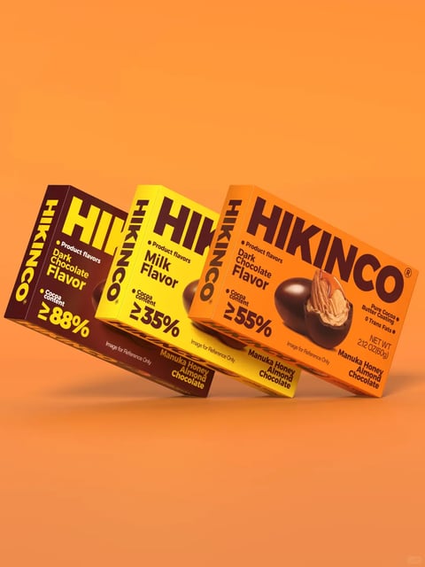 The image displays three rectangular boxes of HIKINCO Manuka Honey Almond Chocolate arranged in a staggered, overlapping composition against a solid, warm orange background. The boxes are oriented diagonally, creating a dynamic, floating effect with soft drop shadows beneath them. The leftmost box is dark brown with bold yellow text, the middle box is bright yellow with dark brown text, and the rightmost box is orange with dark brown text. Each box features the brand name 'HIKINCO' in large, stylized, uppercase letters, along with product details. The rightmost box prominently displays a photographic image of a dark chocolate-coated almond with a bite taken out, revealing the almond inside. The visible text, in English, includes the brand 'HIKINCO', product flavor descriptions like 'Dark Chocolate Flavor' and 'Milk Flavor', cocoa content percentages ('≥88%', '≥35%', '≥55%'), and the product name 'Manuka Honey Almond Chocolate'. Additional text notes 'Pure Cocoa Butter Coating', '0 Trans Fats', and the net weight '2.12 OZ(60g)'. The boxes have a matte finish with crisp, clean edges, and the design uses a bold, modern typographic style. The overall aesthetic is clean, commercial, and product-focused, with a vibrant color palette that differentiates the flavors.