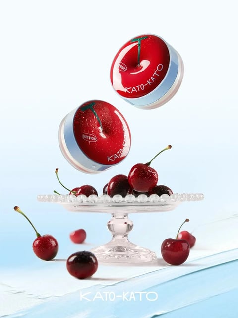 This image is a high-resolution, photorealistic 3D render featuring a clean, bright product advertisement composition. The main subjects are two round, glossy compacts designed to resemble cherries, suspended dynamically in mid-air above a clear glass cake stand holding fresh, dark red cherries. The scene is set against a soft, gradient background transitioning from pale blue at the top to white at the bottom, with a light blue wooden surface in the foreground. The compacts have a shiny red lid mimicking a cherry's skin, complete with a green stem detail, and a silver base; one compact is angled to show its side, while the other is seen more from the top. The glass stand has a textured, beaded rim and a classic pedestal base, holding a small pile of cherries with water droplets, enhancing freshness. Additional cherries are scattered on the surface around the stand, some with stems, creating a sense of natural arrangement. Visible text includes the brand name "KATO-KATO" in white, sans-serif capital letters on the red lids of the compacts and repeated at the bottom center of the image, and the word "REFRESH" in smaller white letters inside a circular emblem on the lids, indicating product branding and a key attribute. The lighting is bright, soft, and diffused, coming from the upper left, casting gentle shadows and creating crisp highlights on the glossy surfaces of the cherries and compacts, emphasizing their smooth, wet texture and vibrant red color. The color language is dominated by saturated cherry reds, cool silver and blue tones, and clean whites, with high contrast between the vivid subjects and the pale background. The composition uses a central, slightly low-angle perspective with floating elements to create depth and dynamism, while the shape language combines the organic, rounded forms of the cherries with the geometric precision of the compacts and glassware. The overall mood is fresh, vibrant, and luxurious, conveying a sense of playful elegance and refreshing appeal.
