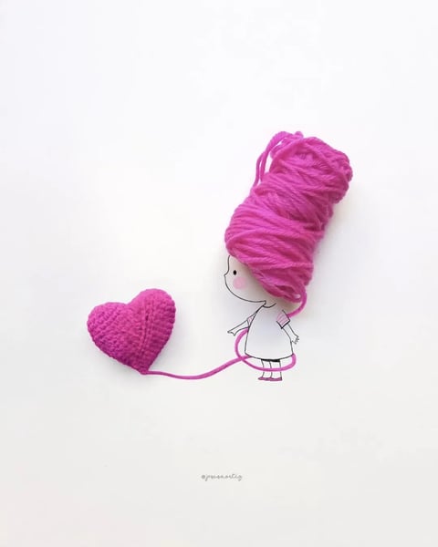 This image is a minimalist, mixed-media illustration combining a simple line drawing with a physical object. The main subject is a small, stylized cartoon girl drawn in thin black ink lines on a plain white background. Her hair is represented by a large, voluminous skein of bright magenta yarn, which cascades down and forms a continuous line that loops around her body like a jump rope, ending in a crocheted magenta heart she appears to be pulling. The composition is asymmetrical, with the girl and her yarn hair positioned on the right, and the heart placed to her left, creating a sense of gentle movement and connection. The visual aesthetic is illustrative and playful, with a flat, graphic quality for the drawing contrasted by the three-dimensional, tactile texture of the yarn. The color palette is starkly limited to the white background, black ink lines, and the dominant, highly saturated magenta of the yarn and heart, creating strong visual contrast. The lighting is soft and even, suggesting a studio or scanned flat-lay setup, with no harsh shadows, which emphasizes the clean, graphic nature of the piece. The texture is a key element: the smooth, matte paper surface contrasts sharply with the soft, fibrous, and intricately stitched texture of the crocheted heart and the loose, twisted strands of the yarn skein. The shape language combines the organic, rounded forms of the yarn and heart with the simple, geometric lines of the girl's figure. The overall mood is whimsical, sweet, and gently romantic, conveying a sense of handmade charm and affectionate connection through the physical link of the yarn. There is no obvious readable text in the main image area, though a very small, faint watermark or signature reading '@jesusaortiz' is visible in the bottom center.