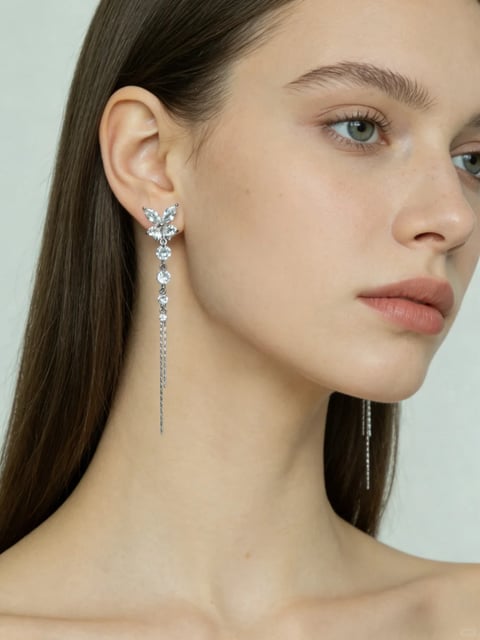 The image is a close-up, photorealistic portrait of a woman's face and neck, framed from the shoulders up against a plain, light gray background. The composition focuses on her left profile, with her head turned slightly toward the viewer, showcasing her sharp jawline, high cheekbone, and long, straight brown hair tucked behind her ear. The primary visual element is a long, dangling earring in her left ear, featuring a silver-toned metal base with a cluster of marquise-cut clear crystals at the top, followed by a vertical line of round crystals and multiple fine, shimmering chain tassels that extend downward. There is no visible text in the image. The lighting is soft and diffused, coming from the front-left, creating gentle highlights on her skin and the facets of the crystals while casting subtle, soft shadows under her jaw and along her neck, conveying a calm and elegant mood. The color palette is muted and natural, dominated by warm skin tones, soft pink lips, cool silver from the earring, and the neutral gray background, with low saturation and balanced contrast. The texture is highly detailed, rendering the smoothness of her skin, the fine strands of her hair, and the sparkling, faceted surfaces of the crystals with a realistic, tactile quality. The shape language combines organic curves of the face and hair with the geometric, angular cuts of the crystals and the straight lines of the dangling chains, creating a balanced contrast between soft and sharp forms.
