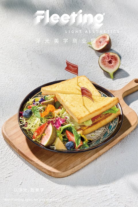 This is a high-angle, photorealistic commercial food photograph centered on a decorative plate holding two golden-brown toasted sandwich halves and a vibrant mixed salad, all presented on a light wooden cutting board against a textured, light gray concrete-like surface. The composition is balanced, with the plate slightly off-center and two halved fresh figs placed in the upper right background to create depth. The visual aesthetic is clean, modern, and appetizing, with a focus on natural textures and fresh ingredients. Visible text includes the large, stylized white word 'Fleeting' at the top, followed by the smaller uppercase phrase 'LIGHT AESTHETICS' and the Chinese characters '浮光美学商业摄影' (Floating Light Aesthetics Commercial Photography) below it. Two small red pennant flags on toothpicks inserted into the sandwiches bear the cursive text 'Four11'. At the bottom left, faint white text reads '以浮光, 致美学' and its English translation 'With Fleeting light, for true aesthetics'. The lighting is bright, soft, and directional from the upper left, casting gentle shadows to the right and creating a fresh, daytime mood. The color palette is dominated by warm, golden tones from the toast, vibrant greens, reds, and purples from the salad and figs, and neutral grays from the background, with high saturation and moderate contrast. Textures are highly detailed, from the crisp, porous surface of the toast and the delicate, veined flesh of the figs to the rough, matte finish of the plate and the fibrous grain of the wooden board. The shape language combines the geometric triangles of the sandwiches with the organic, irregular forms of the salad greens and fruit. The overall mood is inviting, wholesome, and aesthetically refined, emphasizing freshness and quality.