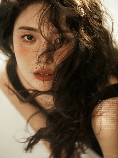 The image is a close-up, photorealistic portrait of a young woman with East Asian features, framed tightly from the shoulders up against a bright, softly blurred background. Her face is the central focus, angled slightly, with dark, wavy hair cascading across her forehead and cheeks, partially obscuring her features and creating a sense of intimate, candid movement. Her skin has a natural, dewy texture with visible freckles across her cheeks and nose, and her lips are parted slightly, painted in a muted, warm terracotta shade. The lighting is soft and directional, appearing to come from the upper left, casting gentle, warm highlights on her hair and the right side of her face while leaving the left side in subtle shadow, enhancing the three-dimensional form and creating a serene, contemplative mood. The color palette is dominated by warm, earthy tones—soft beiges, warm browns in her hair, and the terracotta of her lips—contrasted against the cool, bright white of her off-shoulder top and the background. In the lower right corner, there is small, white, sans-serif text in English that reads, "WHAT DO YOU DO WHEN YOU SEE AN EXCITING SCENE OR AN INTERESTING PERSON ON THE STREET? MANY PEOPLE NOWADAYS WOULD WHIP OUT THEIR CAMERA AND TAKE A SNAPSHOT. AFTER ALL, NOTHING COULD BE EASIER OR MORE FUN THAN STREET PHOTOGRAPHY. SOME PEOPLE, HOWEVER, ARE TAKING IT TO THE NEXT LEVEL—THEY TURN STREET PHOTOGRAPHY INTO ART. BRANDON STANTON, A". The text is overlaid on the image, appearing as a caption or editorial note, and is partially cut off at the bottom. The composition uses a shallow depth of field, keeping the woman's eyes and central facial features in sharp focus while her hair and the background softly blur, drawing the viewer's attention directly to her expressive gaze and the texture of her skin and hair. The overall aesthetic is naturalistic and intimate, with a tactile quality emphasized by the detailed rendering of individual hair strands and skin texture.