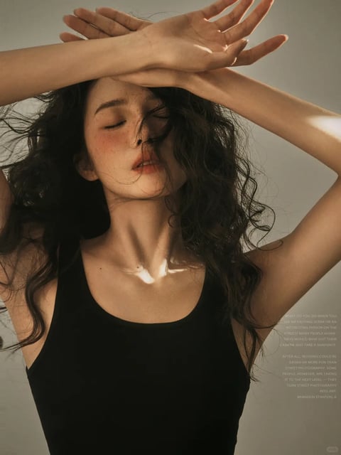 The image is a photorealistic portrait of a young woman with voluminous, dark, curly hair, captured in a close-up, vertical composition. She is positioned centrally, with her arms raised and hands crossed above her head, palms facing inward, creating a dynamic, angular frame around her face. Her eyes are closed, and her head is tilted slightly back, conveying a sense of serenity or introspection. She wears a simple black tank top, and the background is a plain, neutral light gray wall. The visual aesthetic is highly detailed and realistic, with a focus on natural textures. The color language is dominated by warm, earthy tones: her skin has a warm, peachy undertone with visible freckles across her nose and cheeks, her hair is a deep brown with subtle highlights, and the black top provides a strong, low-saturation contrast. The lighting approach is directional and soft, appearing to come from the upper left, casting gentle shadows on the right side of her face and neck while creating bright, warm highlights on her collarbone, shoulder, and the edges of her hair. This lighting enhances the three-dimensionality and adds a calm, golden-hour mood. Texture and material feel are emphasized: the fine, wispy strands of her hair, the smooth yet freckled skin, and the matte fabric of the tank top are all rendered with high tactile detail. Compositionally, the framing is tight, focusing on the upper torso and head, with the raised arms creating leading lines that draw the eye to her face. Shape language is organic, defined by the soft curves of her features and hair contrasted with the sharper angles of her elbows and wrists. The mood and atmosphere are intimate, tranquil, and slightly ethereal. Visible text appears in the lower right corner, printed in a small, white, sans-serif font against the gray background. The readable English text discusses street photography, with phrases like "WHAT DO YOU DO WHEN YOU SEE AN EXCITING SCENE OR AN INTERESTING PERSON ON THE STREET?" and mentions turning street photography into art, signed by "BRANDON STANTON." This text acts as a subtle, graphic overlay, adding a layer of narrative context without distracting from the primary portrait.