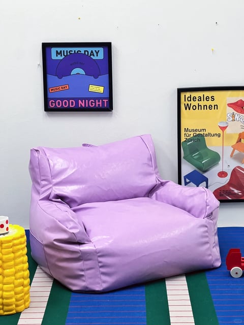 The image presents a vibrant, playful interior scene centered around a large, puffy beanbag chair upholstered in glossy, light purple vinyl, which dominates the lower-middle portion of the frame. The composition is a medium shot against a plain white wall, with the beanbag resting on a striped rug featuring bold bands of royal blue, emerald green, and white with thin red pinstripes. To the left of the chair sits a bright yellow, textured cylindrical side table resembling stacked coins or bubbles, topped with a white mug adorned with red polka dots. On the right foreground, a small red toy car with white wheels is partially visible. The wall behind displays two framed posters: the upper-left one features a graphic design with a purple and blue color scheme, showing a stylized record or sun motif and the words "MUSIC DAY" in bold sans-serif font at the top, "GOOD NIGHT" in pink at the bottom, and smaller text including "MUSIC DAY" on a red label and a yellow box with illegible text. The larger poster on the right has a yellow background and promotes an exhibition titled "Ideales Wohnen" at the "Museum für Gestaltung Zürich," showcasing colorful, modern furniture illustrations like a green chair and a red floor lamp, with text in German. The lighting is bright, even, and diffused, creating soft shadows and highlighting the shiny, smooth texture of the vinyl beanbag and the matte finish of the wall. The overall aesthetic is contemporary and graphic, with a saturated, cheerful color palette dominated by purple, yellow, blue, and red, evoking a fun, youthful, and relaxed atmosphere through its combination of soft, organic shapes in the furniture and clean, geometric lines in the posters and rug.