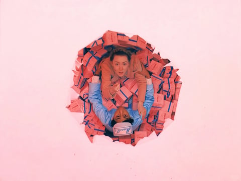 The image presents a striking, surreal composition viewed through a torn, circular hole in a pale pink surface, revealing two figures amidst a chaotic pile of identical pink boxes. The primary visual subject is the interaction between the two individuals: one, a woman with light skin and dark hair pulled back, wearing a light brown jacket, is positioned above, looking directly at the viewer with a surprised or urgent expression, her hand reaching forward as if to grab something. Below her, a man with dark skin, wearing a blue shirt and a white cap with blue script, is upside down, his face also directed towards the viewer with a wide-eyed, startled look, his hand reaching up. They are surrounded by a dense, overlapping mass of rectangular pink boxes, each marked with blue text and graphics, creating a sense of being trapped or overwhelmed. The visible text on the boxes reads 'MODELS' in a simple, sans-serif font, printed in blue, and appears repeatedly across the pile, reinforcing the theme of uniformity or mass production. The man's cap also features blue script, though the specific words are partially obscured and stylized. The visual aesthetic is highly stylized and photorealistic with a painterly touch, characterized by a soft, almost pastel color palette dominated by various shades of pink, from the pale background to the vibrant boxes, contrasted with the cool blue of the man's shirt and the text. The lighting is soft and diffused, appearing to come from above and slightly in front, casting gentle shadows that give depth to the figures and the box pile without creating harsh contrasts, contributing to a dreamlike yet tense atmosphere. Texture and material feel are rendered with precision: the matte paper of the boxes, the fabric of the clothing, and the skin textures of the figures are all distinctly tactile. Compositionally, the circular torn hole acts as a natural frame, drawing the eye directly into the central action and creating a strong sense of depth, with the figures and boxes receding into the space behind the pink surface. The shape language is a mix of the organic, irregular edges of the torn hole and the figures' forms, contrasted with the rigid geometric repetition of the rectangular boxes. The overall mood is one of surreal urgency and confinement, with a high energy level conveyed through the dynamic poses and expressions of the subjects, set against a visually sweet but psychologically pressurized atmosphere.