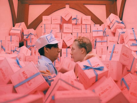This image presents a highly stylized, symmetrical composition centered on a close, intimate profile gaze between a young man and woman, who are surrounded by a chaotic, towering avalanche of pale pink pastry boxes that fill the entire frame. The visual aesthetic is distinctly cinematic and meticulously art-directed, featuring a soft, painterly photorealism with a dreamlike quality. The color language is dominated by a cohesive, low-contrast pastel palette of soft pinks, creams, and light blues, with the warm pink hue of the boxes and background infusing the entire scene with a gentle, monochromatic warmth, while the cool blue of the ribbons and the man's uniform provides a subtle, complementary accent. The lighting is soft, diffused, and ambient, seemingly emanating from all directions to create a flat, even illumination that minimizes harsh shadows and enhances the ethereal, stage-like atmosphere. The texture and material feel are rendered with a tactile smoothness; the boxes appear to be made of matte cardboard with a slight sheen, tied with satin-like blue ribbons that catch soft highlights, while the clothing and hair have a soft, tangible quality. The composition is dense and claustrophobic yet balanced, with the two figures framed centrally amidst the chaotic, asymmetrical stacking of the geometric boxes, creating a sense of depth through overlapping forms. The shape language is a contrast between the rigid, repetitive geometry of the cubic boxes and the soft, organic curves of the figures' faces and hair. The overall mood is one of whimsical melancholy, intimate tension, and surreal romance, conveying a quiet, suspended moment within a visually overwhelming environment. The visible text includes the word 'MENDLS' printed in a classic, serif font on the sides of numerous pink boxes, along with smaller decorative text like 'CHOCOLATES' and 'VIENNESE', establishing the setting as a patisserie or confectionery shop.