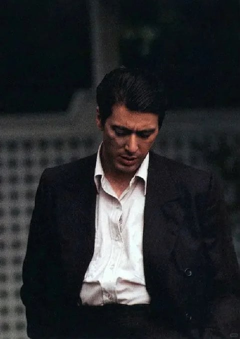 The image features a medium shot of a man, Alain Delon, positioned centrally within the frame, looking downward with a somber, introspective expression. The composition is tightly focused on his upper body and head, set against a blurred, indistinct background that suggests an outdoor or architectural setting with a patterned fence or wall, rendered with a shallow depth of field that isolates the subject. The visual aesthetic is deeply cinematic and photorealistic, with a grainy, filmic texture reminiscent of classic 1960s cinema. The color language is characterized by a desaturated, moody palette dominated by deep blacks, charcoal greys, and stark whites, with cool undertones that enhance the somber atmosphere; the contrast is high, with the dark suit jacket sharply defining against the white shirt and pale skin. Lighting appears to be soft and diffuse, coming from above and slightly to the front, casting gentle shadows under the brow and chin that emphasize the subject's sharp facial structure and contemplative mood without harshness. Texture and material are conveyed through the matte wool of the suit jacket, the crisp cotton of the unbuttoned dress shirt, and the fine strands of his slicked-back dark hair, all rendered with a tactile, film-grain quality. There is no visible or readable text in the image. The mood is melancholic, intense, and brooding, with a heavy visual weight centered on the subject's downward gaze and the overall dark tonal scheme.