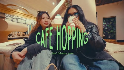 The image captures a candid indoor scene with two young women sitting side-by-side on a cream-colored, tufted sofa in a modern, stylish café or lounge, shot from a medium close-up, slightly low-angle perspective. The overall composition is centered on the two figures, framed by warm architectural elements and soft lighting. The visual aesthetic is photorealistic with a cinematic, slightly warm-toned color grade, emphasizing rich textures and a cozy atmosphere. Dominant colors include the deep black of leather and fabric, soft heather gray denim, warm amber from ambient lighting and wood tones, and vibrant lime green from the prominent text overlay. The lighting is soft and directional, appearing to come from overhead recessed lights and possibly a window to the left, creating gentle highlights on faces, hair, and surfaces, with soft shadows that enhance depth without harsh contrast. Textures are rendered with tactile detail: the supple, slightly shiny grain of a black leather jacket, the matte softness of a gray knit sweater, the coarse weave of light-wash denim jeans, and the plush, velvety surface of the sofa. The composition uses a shallow depth of field, keeping the subjects sharp while the background—featuring blurred clothing racks, a graffiti-style mural, and a door with leafy wallpaper—recedes softly. Shape language is a mix of organic curves from the figures and sofa against geometric lines from the architecture and shelving. Large, bold, sans-serif text in a vibrant lime green is overlaid centrally, reading "CAFE HOPPING" in all caps, with smaller, cursive text below stating "feat. bianca gan", playing a dominant graphic role that contrasts with the realistic scene. The mood is relaxed, youthful, and social, conveying a casual, stylish outing with a warm, inviting, and slightly trendy atmosphere.