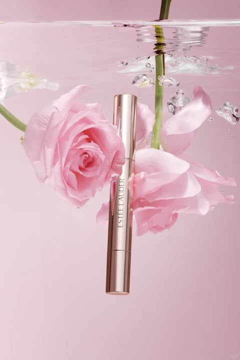 The image features a sleek, cylindrical Estée Lauder cosmetic product, likely a mascara or eyeliner tube, centered vertically against a soft, gradient pink background. The metallic, rose-gold-toned tube is suspended in clear water, surrounded by floating pink roses and petals that drift around it, with small air bubbles clinging to the stems and petals, creating a serene, submerged scene. The composition is balanced and elegant, with the tube as the focal point, flanked by the lush floral elements that extend from the top left and right. The text 'ESTÉE LAUDER' is printed vertically in a clean, sans-serif, dark gray font along the side of the tube, clearly identifying the brand. The tube has a smooth, highly reflective metallic surface with a glossy finish, showing subtle highlights and reflections, and is constructed with a subtle seam indicating a cap and body. Its form is a simple, elongated cylinder with uniform proportions, suggesting a minimalist and luxurious style. The surrounding water and flowers imply a context of freshness, purity, and natural beauty, with the lighting soft and diffused, enhancing the delicate, romantic mood.