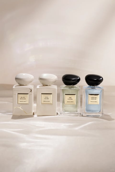 The image displays a row of four Armani Privé perfume bottles arranged side-by-side on a smooth, neutral-toned surface, likely a table or shelf, against a softly lit, beige or off-white background. The composition is centered and horizontal, with the bottles evenly spaced. The two bottles on the left feature opaque, creamy white or off-white rectangular glass bodies with subtle gold or grey veining, topped with smooth, rounded, white, pebble-like caps. The two bottles on the right are made of clear glass, revealing pale green and light blue liquids inside, and are topped with contrasting smooth, black, pebble-like caps. Visible text on each bottle is printed on a small, centered, rectangular label with a gold border. From left to right, the labels read 'BLANC KOGANE', 'OUD NACRÉ', 'THÉ YULONG', and 'VÉTIVER D'HIVER', all with 'ARMANI / PRIVÉ' written below in smaller text. The lighting is soft and diffused, coming from the upper left, creating gentle highlights on the rounded caps and subtle shadows to the right of each bottle, emphasizing their three-dimensional form and the smooth, polished texture of the glass and caps. The overall aesthetic is minimalist, elegant, and photorealistic, with a focus on clean lines, organic shapes in the caps, and a luxurious, serene atmosphere conveyed by the muted color palette of whites, creams, blacks, and pale liquids.