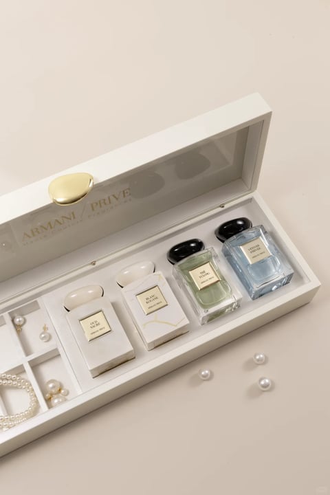 This is a photorealistic, high-key product photograph featuring a luxurious, off-white, rectangular gift box with a transparent lid, angled open to reveal its contents. The box is the central subject, positioned diagonally from the lower-left to the upper-right against a smooth, uniform, light beige background. Inside, nestled in molded white compartments, are four small, rectangular perfume bottles from the ARMANI/PRIVÉ line, each with a distinct cap color and liquid hue: from left to right, a white marble-patterned bottle with a white cap labeled 'OUD NACRÉ', a white bottle with gold veining and a white cap labeled 'BLANC KANKAN', a clear bottle with pale green liquid and a black cap labeled 'THE YLONG', and a clear bottle with pale blue liquid and a black cap labeled 'VETIVER D'HIVER'. To the left of the perfumes, separate compartments hold delicate pearl jewelry: a pair of pearl drop earrings and a small pearl bracelet. A few loose, individual pearls are scattered on the background surface near the bottom right corner. The visible text, rendered in a clean, gold serif font, appears on the inside of the lid as 'ARMANI / PRIVE' with 'Haute Couture Fragrances' in smaller script beneath it, and on each perfume bottle's label, identifying the scent name and brand. The lighting is soft and diffused from an overhead source, creating gentle, even illumination with minimal harsh shadows, highlighting the glossy surfaces of the bottles and the matte texture of the box. The composition uses a shallow depth of field, with the perfumes and jewelry in sharp focus while the background and box edges soften slightly, creating a sense of refined elegance and luxury. The color palette is muted and sophisticated, dominated by creamy whites, soft beiges, and gold accents, with subtle pops of pale green, blue, and the black caps for contrast. The overall mood is serene, opulent, and meticulously arranged, emphasizing the premium quality and aesthetic appeal of the products.