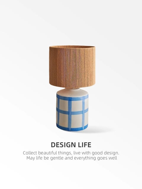The image presents a single, stylized table lamp centered against a plain, off-white background in a studio product shot. The lamp features a cylindrical ceramic base in a soft cream or beige tone, decorated with a hand-painted blue grid pattern where the lines vary slightly in opacity and thickness, giving it an artisanal, imperfect feel. Atop the base sits a textured, drum-shaped lampshade made of a natural, straw-like material with visible vertical fibers in warm, golden-brown tones. The text "DESIGN LIFE" appears in bold, dark gray sans-serif font directly below the lamp, followed by a smaller, lighter gray subtitle that reads: "Collect beautiful things, live with good design. May life be gentle and everything goes well". The lighting is soft and diffuse, coming from the upper left, casting a gentle shadow to the right and emphasizing the tactile qualities of the lampshade's fibers and the smooth ceramic glaze. The overall aesthetic is minimalist and organic, blending geometric shapes with natural textures to evoke a sense of calm, mindful living.