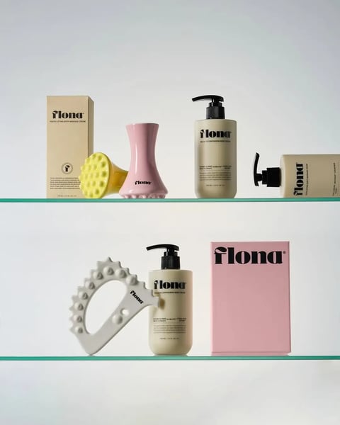 This is a clean, minimalist studio product photography shot featuring the Flona brand's body care line, arranged across two floating glass shelves against a plain, light grey background. The composition is balanced and airy, showcasing a mix of skincare products (beige pump bottles and cardboard boxes) and accompanying massage tools (a pink vase-shaped tool, a yellow mushroom-shaped tool, and a white ring-shaped tool) with distinct, rounded geometric forms. The color palette is soft and muted, dominated by warm beige, gentle pastel pink, pale yellow, and cool off-white, accented by black pump mechanisms and text. The lighting is bright, even, and highly diffused, creating soft, subtle shadows that emphasize the smooth, matte plastic and paper textures without creating harsh contrast, contributing to a serene and clinical atmosphere. The visual aesthetic is modern, sleek, and product-focused, with a 3D-rendered or high-end photographic quality that highlights the tactile, ergonomic shapes of the tools. Readable text is visible on all product packaging and tools, consistently featuring the brand name 'flona' in a bold, modern serif font, along with product descriptions in smaller sans-serif type such as 'YOUTH LIFTING BODY MASSAGE CREAM' and 'CELLULITE CONTOURING BODY CREAM', primarily in English. The spatial arrangement is deliberate, with objects placed at varying depths on the shelves to create visual interest while maintaining a sense of organized, spacious display.