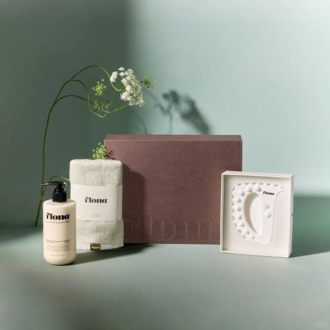 This image presents a serene, minimalist lifestyle product arrangement set against a smooth, muted sage green surface and background, captured in a photorealistic style with soft, diffused lighting. The composition is balanced and centered, featuring a curated collection of wellness items. A large, rectangular gift box with a textured, mauve-brown surface and debossed 'flona' text serves as the central backdrop. To its left stands a cylindrical bottle of 'flona' hand wash in a matte beige container with a black pump, and beside it, a tightly rolled, off-white linen towel secured by a paper band also bearing the 'flona' brand. To the right of the box, a white scalloped scalp massager is presented within a shallow, light-colored tray. A delicate stem of white flowering herbs, possibly Queen Anne's lace, arches gracefully from behind the towel, adding an organic, natural element that casts a soft, intricate shadow on the background wall. The color palette is dominated by earthy neutrals—mauve, beige, off-white, and sage green—creating a low-contrast, calming atmosphere. Text is clearly visible on multiple items: the brand name 'flona' appears in a clean, lowercase serif font on the hand wash bottle, the towel band, the gift box (debossed), and the scalp massager, establishing brand consistency. The lighting is soft and directional from the upper left, creating gentle gradients and subtle shadows that enhance the tactile qualities of the textured box, the linen towel, and the smooth matte surfaces. The overall mood is one of understated elegance, purity, and mindful self-care, with a strong emphasis on natural materials and a cohesive, branded aesthetic.