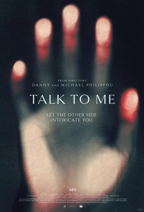 This is a horror movie poster dominated by a large, out-of-focus, disembodied hand with a pale, slightly grayish skin tone. The hand is positioned palm-forward, filling most of the vertical composition, with its fingers spread wide. The tips of the fingers are the most distinct element, glowing with a vivid, luminous red-orange light that fades into a darker, blurred red towards the base of the fingernails, creating a stark contrast against the grainy, dark, near-black background. Centered over the palm of the hand is white, serif text. At the top, smaller text reads 'FROM DIRECTORS DANNY AND MICHAEL PHILIPPOU'. Below this, the main title 'TALK TO ME' is displayed in a large, elegant, all-caps font. Underneath the title, a smaller tagline states 'LET THE OTHER SIDE INTOXICATE YOU'. At the very bottom, in fine print, are production credits including the A24 logo and other text listing producers, writers, and cast. The focus is sharply on the glowing red fingertips, while the rest of the hand and the background are heavily blurred and grainy, emphasizing the supernatural, eerie quality of the central subject. The overall mood is unsettling and mysterious, with a dark, moody color palette of blacks, deep grays, and the striking red-orange of the fingertips.