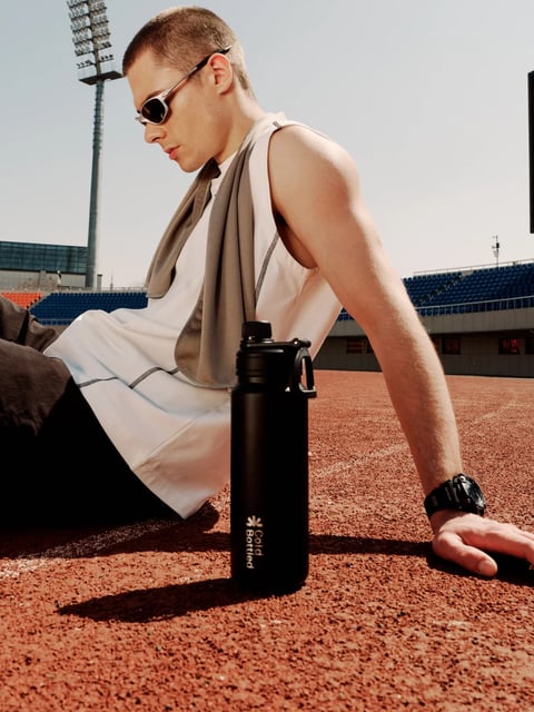 The image presents a low-angle, photorealistic composition centered on a young man resting on a reddish-brown, granular running track, with his body angled diagonally across the frame from the lower-left to mid-right. He wears a sleeveless, off-white athletic tank top with visible black stitching, dark trousers, a grey towel draped around his neck, and silver-framed sunglasses, with his left arm bracing his weight on the ground and his right hand resting near his knee. The scene is set in an outdoor stadium with rows of blue and orange seating visible in the mid-ground, under a bright, clear sky. A tall stadium light pole rises in the upper-left background. The dominant color palette features warm terracotta tones from the track, muted neutrals from the clothing, and cool blues from the stadium seats, with high saturation and strong contrast created by harsh, directional sunlight that casts sharp, dark shadows from the subject and the water bottle onto the track. The lighting is hard and unfiltered, suggesting midday sun, creating bright highlights on the subject's skin and clothing and deep, defined shadows that emphasize the granular texture of the track surface. The primary object in sharp focus is a matte black water bottle with a rigid plastic lid and handle, positioned upright in the foreground slightly right of center; it features vertical, sans-serif text in white reading "Cold" above "Bottled" alongside a small logo, which is the only clear readable text in the image. The texture of the track is rough and pebbly, contrasting with the smooth matte finish of the bottle and the soft fabric of the towel and tank top. The composition uses a shallow depth of field, keeping the subject and bottle in sharp focus while the stadium background is softly blurred, and the shape language combines the organic forms of the human figure with the geometric lines of the stadium architecture and the cylindrical, industrial design of the bottle. The overall mood is one of athletic rest and recovery, with a focused, intense energy conveyed by the harsh lighting and the subject's relaxed yet powerful posture.