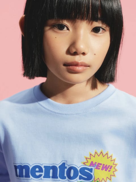 The image presents a close-up, photorealistic portrait of a young woman with a straight, blunt-cut black bob hairstyle with bangs, framed from the shoulders up against a solid, soft pink background. She faces the camera directly with a neutral, calm expression, her dark eyes looking straight ahead. She wears a light blue, heathered cotton t-shirt with a ribbed crew neckline. Visible text on the shirt includes the word "mentos" in a bold, rounded, dark blue sans-serif font with a white outline, positioned horizontally across the chest. To the right of this text is a yellow, spiky, starburst-shaped graphic containing the word "NEW!" in a bold, pink, all-caps sans-serif font. The lighting is bright and directional from the upper left, creating soft, defined shadows on the right side of her face, neck, and under her chin, with a high-contrast, clean quality that suggests studio lighting. The color palette is dominated by the cool pastel blue of the shirt and the warm pastel pink background, creating a complementary, graphic, and youthful contrast. The texture of the t-shirt is rendered with fine detail, showing the soft, slightly fuzzy knit of the fabric. The composition is centered and tightly cropped, focusing attention on the subject's face and the branded graphic on her shirt, with a shallow depth of field that keeps her features sharp. The shape language is characterized by the sharp, geometric lines of the bob haircut and the spiky starburst graphic, contrasting with the organic, soft curves of her facial features. The overall mood is cool, serene, and stylistically modern, with a clean, high-fashion, commercial aesthetic.