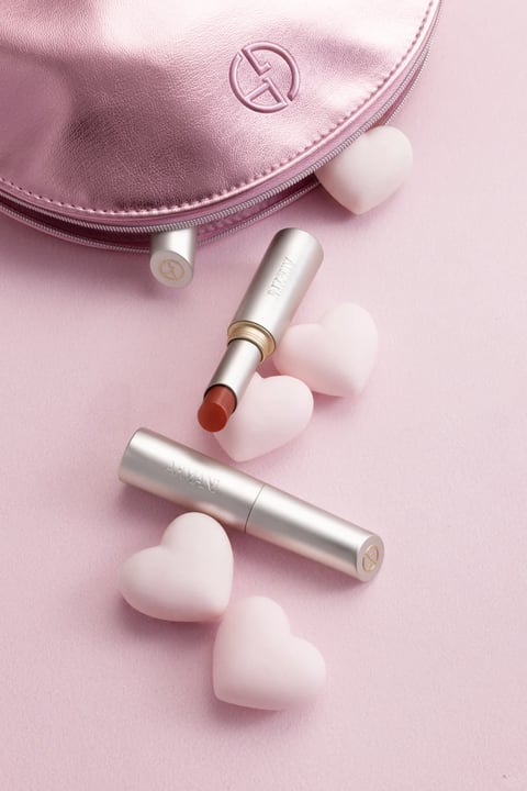 This image presents a soft, romantic still-life composition centered on two metallic silver Giorgio Armani lipstick tubes, one open to reveal a warm terracotta shade, and a matching metallic pink satin makeup pouch with a prominent embossed 'GA' logo and a silver zipper pull. The scene is arranged on a seamless light pink matte surface, with several smooth, heart-shaped pastel pink confections scattered around the products, creating a cohesive Valentine's or gift-themed aesthetic. The visual style is highly photorealistic, with a focus on luxurious material rendering. The color palette is monochromatic and dominated by soft pinks, complemented by the cool metallic silver of the packaging and the single warm pop of color from the exposed lipstick bullet, maintaining low saturation and high tonal harmony. Lighting is soft, diffused, and directional from the upper left, casting gentle, blurred shadows that enhance the three-dimensional form of the objects without creating harsh contrast, conveying a serene and gentle mood. Textures are meticulously rendered: the lipstick tubes exhibit a brushed, anodized aluminum finish with fine engraved serif text reading 'ARMANI', while the pouch displays a soft, pebbled leather-like grain with a subtle satin sheen on the metallic pink surface; the heart shapes appear smooth, matte, and fondant-like. Compositionally, the objects are arranged in a loose, diagonal flow from the upper left pouch down toward the lower right, utilizing shallow depth of field to keep the foreground lipstick sharp while allowing the pouch in the background to soften slightly. Shape language combines the strong cylindrical geometry of the lipsticks with the organic, soft curves of the heart shapes and the rounded pouch, with clean, precise edges on the manufactured items. The overall atmosphere is one of delicate luxury, sweetness, and romantic elegance, with low visual weight and high tactile appeal.