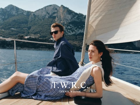 This image is a high-fashion, photorealistic editorial shot set on a wooden sailboat deck, framed by a vast, deep blue sea and rugged coastal mountains under a clear sky. The composition centers on a man and woman in sophisticated, relaxed summer attire; the man, positioned slightly behind, wears a dark blue textured quarter-zip sweater and sunglasses, gazing off-camera with a composed expression, while the woman reclines in the foreground, dressed in a blue and white micro-check sleeveless dress with a cream sweater draped over her shoulders, her hair caught in a light breeze. Visible text is overlaid in the lower center: "T.W.R.C." in a bold, white serif font, with "TEENIE WEENIE" in a smaller, matching font beneath it, serving as a branding logo. The visual aesthetic is polished and aspirational, with a color palette dominated by cool, saturated blues—from the navy sweater to the ocean—complemented by warm wooden deck tones and crisp whites, creating a high-contrast, nautical-inspired look. Lighting is bright and directional, suggesting midday sun with soft, diffused shadows that enhance the textures of the knit sweater, the woven dress fabric, and the smooth wood grain. The composition uses a shallow depth of field to keep the subjects sharp against the slightly blurred mountain backdrop, with dynamic diagonal lines from the sail and railing guiding the eye across the frame. Shapes are a mix of organic forms (the figures, hair, mountains) and geometric elements (the boat's railing, sail edges), while the mood is one of serene, elegant leisure, evoking a sense of affluent, relaxed sophistication.