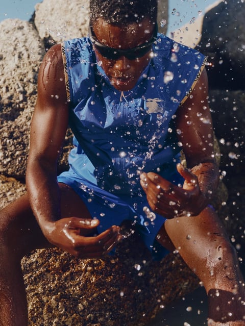 The image is a photorealistic, high-angle shot capturing a Black male athlete seated on rugged, dark, wet rocks, likely in a coastal or outdoor training setting. The subject, wearing a vibrant royal blue sleeveless athletic top with gold trim and matching shorts, is framed in a dynamic composition where water droplets are suspended in mid-air around him, creating a sense of motion and intensity as he looks downward with his hands cupped as if catching or interacting with the splashing water. There is no obvious readable text in the image. The lighting is high-contrast with a hard, directional quality, likely from a midday sun, creating strong, bright highlights on the wet skin, the reflective water droplets, and the textured rocks, while casting deep, cool-toned shadows. The color palette is dominated by the vibrant blue of the athletic wear, contrasting sharply with the warm, earthy tones of the athlete's skin and the muted, dark greys and browns of the damp rock surfaces. The textures are highly detailed, emphasizing the slickness of the wet skin, the individual, sparkling water droplets, and the rough, porous surface of the rocks. The composition uses a shallow depth of field, keeping the athlete and the immediate water spray in sharp focus while the background rocks blur slightly, enhancing the sense of immediacy and focused energy. The overall mood is one of intense focus, athleticism, and raw, energetic action, conveyed through the dynamic water elements and the subject's concentrated posture.