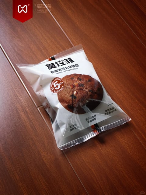 This image features a single, sealed plastic package of bread resting diagonally on a dark, polished wooden surface with visible grain lines. The overall visual aesthetic is photorealistic, focusing on product photography with a clean, commercial feel. The dominant color language is warm, with the deep reddish-brown tones of the wood providing a rich background that contrasts with the predominantly white and silver packaging. The lighting approach uses soft, directional light from the upper left, casting gentle shadows to the right and below the package, highlighting the crinkles in the plastic and the sheen of the wooden floor, creating a warm and inviting mood. The texture and material feel are distinct: the smooth, slightly reflective plastic of the packaging contrasts with the matte, textured print of the bread illustration on it and the glossy, linear grain of the wooden floor. The composition follows a diagonal arrangement, placing the product centrally but at an angle to create dynamic visual interest, with shallow depth of field keeping the package in sharp focus. The shape language is a mix of the organic, rounded form of the bread illustration and the irregular, crumpled geometric shape of the sealed package. The mood and atmosphere are casual and appetizing, suggesting a convenient snack. Visible text includes the large brand name '莫拉菲' and product name '香橙巧克力味欧包' (Orange Chocolate Bread) in Chinese, with smaller English text 'ORANGE CHOCOLATE BREAD'. A prominent red circular badge states 'FREEZE TO FRESH 5min' and '核心冷冻鲜味'. Other text specifies '72小时液种发酵' (72-hour liquid fermentation), '黑巧克力味' (Dark Chocolate Flavor), and '净含量:85克' (Net Weight: 85g), all playing a key informational and branding role. In the upper left corner, a red banner logo and text '© WWW.NOE.HK NOE FACTORY BY EASON ZHENG MAY 5, 2026' are superimposed as a watermark.