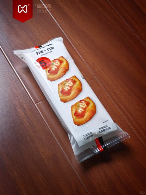 The image presents a high-angle, close-up shot of a single package of Danish pastry resting diagonally on a dark wooden floor with visible grain patterns. The main subject is a rectangular, sealed plastic packet with a clean white front panel displaying three identical, golden-brown, square-shaped Danish pastries arranged vertically. Each pastry features a shiny, glazed surface with a central red fruit filling and a delicate zigzag of white icing. The composition is asymmetrical, with the packet placed from the upper left to the lower right, creating a dynamic diagonal line against the parallel lines of the wooden floorboards. The lighting is soft and directional, coming from the upper left, casting a subtle shadow to the right and below the packet, which enhances its three-dimensional form and the glossy texture of the pastries. The visual aesthetic is photorealistic, with a focus on clean product photography; the rendering is sharp, emphasizing the flaky, layered texture of the pastry crust and the reflective quality of the glaze. The color palette is dominated by the warm golden-brown and amber tones of the pastries and the rich, dark brown of the wood, contrasted against the neutral white of the packaging. The text on the package is primarily in Chinese characters, with some English; at the top, '丹麦一口酥' and 'DANISH PASTRY' identify the product, while a red circular badge with the number '5' and text indicating '5层起酥' (5-layer puff pastry) sits beside the top pastry. Further down, smaller text reads '二次发酵' (secondary fermentation) and '72层开酥' (72-layer lamination), with product details like '调理面包' (prepared bread) and '净含量:80克' (net weight: 80g) near the bottom. The overall mood is commercial and appetizing, with a focus on product appeal and quality, conveyed through the precise, well-lit presentation and the emphasis on textual claims of artisanal technique.