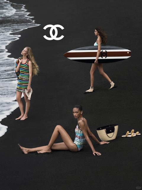 The image features a high-fashion, editorial-style photograph set on a stark, black volcanic sand beach, with gentle white ocean waves lapping at the shore on the left. The composition is structured with three models in different poses and attire, creating a dynamic triangular arrangement against the high-contrast backdrop. The central visual element is the iconic Chanel logo—two interlocking white 'C's—prominently placed in the upper left quadrant. A model on the left stands barefoot at the water's edge, wearing a vibrant, horizontally striped knitted tank dress in multiple colors and holding a small beige pouch. In the upper right, a second model walks across the sand, carrying a surfboard with brown and white stripes and a black Chanel logo, wearing a light blue bikini top and beige ankle boots with a small interlocking 'C' logo. Seated in the foreground, a third model wears a blue and green patterned one-piece swimsuit, looking directly at the viewer; beside her on the sand rests a large woven tote bag with a black leather top and chain strap, and a pair of beige slide sandals with fuzzy accents. The visual aesthetic is photorealistic and highly stylized, resembling a luxury campaign. The color language is defined by the dramatic contrast between the deep, neutral black of the sand and the saturated, varied hues of the clothing and accessories. The lighting is soft and diffused, likely overcast daylight, creating even illumination with minimal harsh shadows, which emphasizes the textures of the materials. Texture and material feel are highlighted through the granular matte surface of the sand, the knitted weave of the dress, the smooth polished surface of the surfboard, the natural straw of the bag, and the plush suede of the footwear. The composition uses a slightly elevated perspective and spatial arrangement that balances the figures across the frame, with the ocean line adding a diagonal element. Shape language combines organic forms of the human figures and waves with the sharp geometric lines of the logo, surfboard, and accessories. The overall mood is one of sophisticated, aspirational leisure with a cool, serene atmosphere, elevated by the minimalist yet luxurious styling.