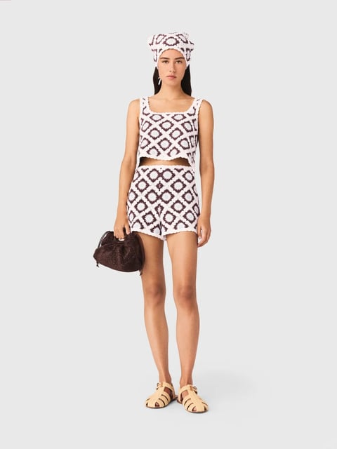 The image presents a full-body, studio-lit portrait of a female model centered against a seamless, neutral light gray background, showcasing a coordinated crochet fashion ensemble. The model stands in a relaxed, forward-facing pose, holding a dark brown textured handbag in her right hand. She wears a two-piece crochet outfit consisting of a cropped, square-neck tank top and matching high-waisted shorts, both featuring a bold geometric diamond pattern in cream and deep burgundy. Her look is completed with a matching crochet headwrap in the same colorway, simple gold hoop earrings, and beige leather slide sandals with cut-out details. The visual aesthetic is photorealistic with clean, commercial styling, emphasizing the tactile quality of the handcrafted textile. The color palette is grounded in warm neutrals—cream, beige, and rich brown—creating a cohesive, earthy, and summery feel with high contrast between the cream and burgundy patterns. Lighting is soft, even, and frontal, minimizing harsh shadows to clearly display the garment's texture and construction. The texture and material feel are highly prominent, with the looped, knitted surface of the crochet work providing a distinct, tactile, and handmade quality against the smooth skin of the model and the suede-like finish of the handbag. Compositionally, the subject is centrally framed with ample negative space, focusing all attention on the outfit's silhouette and pattern. Shape language is defined by the repetitive geometric diamond motifs of the crochet, which contrast with the organic curves of the model's body and the rounded form of the handbag. The overall mood is fresh, relaxed, and stylish, conveying a sense of casual, artisanal summer fashion.