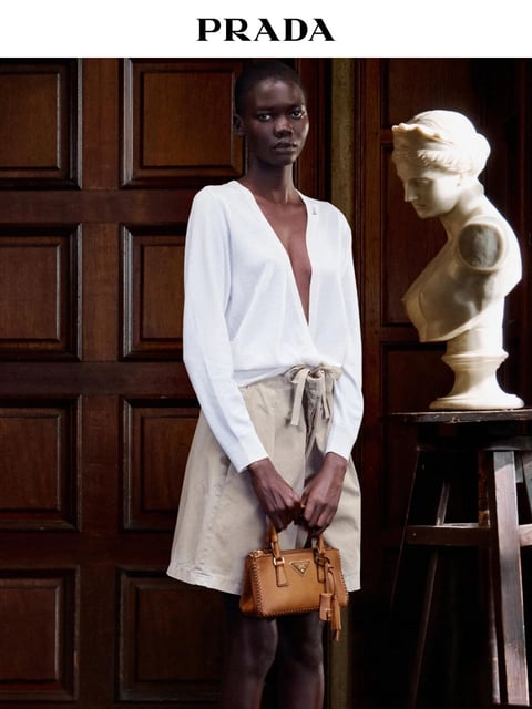 This is a photorealistic fashion photograph featuring a dark-skinned female model positioned centrally against a rich, dark brown wooden paneled wall. She wears a minimalist ensemble: a loose, white, V-neck long-sleeved top with a deep plunging neckline and a wrap-style, knee-length skirt in a natural beige linen fabric, secured with a tied waist sash. In her hands, she holds a structured, tan leather handbag with visible topstitching and gold-tone hardware. To her right, placed on a dark wooden stool, is a classical white plaster bust of a female figure, creating a striking contrast in form and material. The text 'PRADA' appears in bold, black, serif capital letters at the top center of the image against a white bar, identifying the brand. The lighting is soft and directional, coming from the left, casting gentle shadows that define the model's features, the fabric folds, and the classical contours of the bust, evoking a calm, studio-like atmosphere. The composition is balanced and vertical, with the model and sculpture occupying the middle ground, creating a dialogue between contemporary fashion and classical art. The color palette is muted and sophisticated, dominated by whites, beiges, and deep browns, with the warm tan of the bag providing a subtle accent. The overall aesthetic is clean, elegant, and editorial.