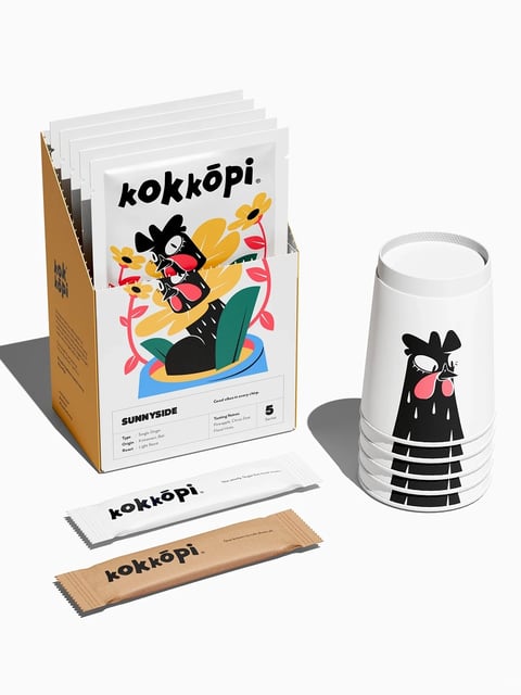 The image displays a cohesive product packaging and accessory set for a coffee brand named 'kokkōpi', presented against a clean, off-white studio background with soft, directional lighting from the upper left casting subtle shadows. The central composition features a rectangular cardboard box with a white front panel and orange side panels, containing multiple individual white coffee sachets. The sachets and box are adorned with a distinctive, playful illustrative style characterized by bold black outlines, flat color fills, and a whimsical, slightly abstract bird-like character with prominent pink cheeks and expressive eyes. To the right stands a stack of white paper cups, each printed with a simplified, elongated version of the same black character. In the foreground, two loose sachets—one white and one brown kraft paper—lie flat, showcasing the brand's logo and minimal text. The overall visual aesthetic is a clean, modern mix of illustrative charm and minimalist product photography, rendered with crisp, high-definition clarity. The color language is dominated by a high-contrast palette of pure white, deep black, and a warm, muted orange on the box sides, accented by vibrant pops of coral pink, sunny yellow, and teal within the illustrative elements. The lighting is soft yet defined, creating a bright, approachable mood that emphasizes the product's tangible qualities. Text is clearly visible throughout: the brand name 'kokkōpi' in a custom, rounded, lowercase sans-serif font appears on the box, sachets, and cups; product details on the box include 'SUNNYSIDE', 'Type: Single-Origin', 'Origin: Kintamani, Bali', 'Roast: Light Roast', 'Tasting Notes: Pineapple, Citrus Zest, Floral Hints', and the phrase 'Good vibes in every sip'; smaller text on the sachets reads 'Use mostly Sugar but trust taste' and 'One bitten to rule them all.' The composition is balanced and spatially arranged to guide the eye from the upright box to the stack of cups and down to the foreground sachets, creating a sense of a complete, ready-to-use kit. The shape language combines the strict geometric forms of the rectangular box, cylindrical cups, and linear sachets with the fluid, organic shapes of the illustrated character and floral motifs, all rendered with sharp, clean edges. The overall mood is cheerful, artisanal, and inviting, suggesting a premium yet fun coffee experience.