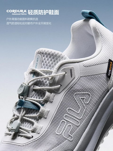 The image is a high-resolution, photorealistic product photograph focusing on the upper portion of a light gray athletic shoe, presented against a soft, gradient blue-gray background. The shoe is angled slightly upward from the bottom left to the top right, showcasing its intricate construction. The primary visual elements are the layered textures: a breathable, honeycomb-patterned mesh forms the main body, overlaid with smooth, matte synthetic panels and reinforced fabric sections. A distinctive quick-lace system with light gray toggles and cords replaces traditional laces, threaded through fabric loops. A small, textured rubber piece shaped like a stylized 'F' adorns the tongue. The overall visual aesthetic is clean, modern, and technical, emphasizing functionality. The color palette is dominated by cool, muted tones of light gray, off-white, and cool blue, with a single pop of vibrant teal on the heel pull-tab and a small tongue loop, creating a low-saturation, high-key look with subtle contrast. The lighting is soft and diffused, coming from the upper left, casting gentle shadows that define the shoe's contours and textures without harshness, conveying a calm, premium mood. Textures are rendered with high detail, from the porous, tactile feel of the mesh to the smooth, slightly granular surface of the synthetic overlays and the fine weave of the fabric loops, suggesting durability and breathability. The composition uses a tight, diagonal crop that fills the frame, creating dynamic energy and depth through the layered materials. Shape language combines organic curves in the shoe's silhouette with precise geometric patterns in the mesh and panels. Visible text includes "CORDURA Advanced Fabrics" in white sans-serif font at the top left, with smaller Chinese characters below describing the material's properties; "FILA VOLLE ATR" is vertically printed on a tongue tag; and a black "CORDURA" label is stitched near the heel. The overall mood is one of advanced, lightweight performance and urban readiness.