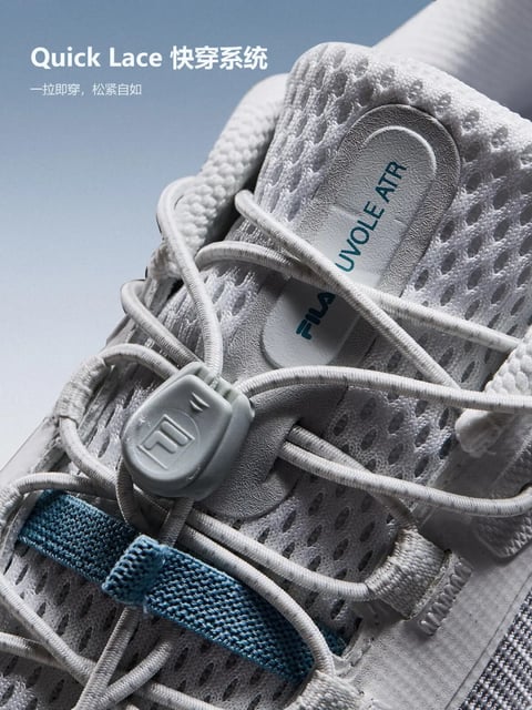 The image is a photorealistic, close-up studio shot of a light grey sneaker, focusing on the upper tongue and lacing area against a soft, gradient blue-grey background. The visual aesthetic is clean, modern, and product-focused, with high detail and sharp focus on the textures. The color language is dominated by cool, neutral tones: the sneaker features multiple shades of grey, from the light mesh to the darker grey suede-like overlays and lace toggle, accented by a single horizontal strap in a muted, dusty blue. Text is present and plays a key branding and informational role. At the top left, white sans-serif text reads 'Quick Lace 快穿系统' and '一拉即穿，松紧自如', describing the lacing feature in both English and Chinese. On the sneaker's tongue, a light grey leather-like patch is printed with the vertical text 'UVOLE ATR' in teal, and below it, the 'FIL'A' logo appears in the same teal. The lighting approach is soft and directional, coming from the upper left, creating gentle highlights on the mesh and smooth gradients on the suede, conveying a clean, commercial mood. The texture and material feel are highly tactile: the upper is a breathable, open-weave mesh with a porous texture, contrasted by smooth, matte suede overlays and the soft, cord-like quality of the elastic laces. The composition is tightly framed, using a shallow depth of field to keep the lacing system and branding in sharp focus while the background and shoe edges softly blur, creating depth. Shape language combines organic, flowing curves from the laces with the geometric precision of the mesh holes and the angular, branded lace toggle. The overall mood and atmosphere are technical, calm, and premium, emphasizing functional design and material quality with a restrained, cool-toned energy.