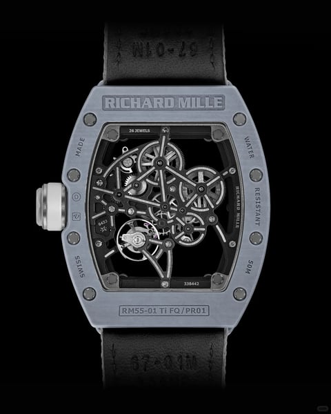 The image presents a high-contrast, photorealistic studio shot of the case back of a Richard Mille RM55-01 Ti FQ/PR01 automatic tourbillon wristwatch, centered against a solid black background. The composition is tightly framed and symmetrical, focusing entirely on the tonneau-shaped titanium case and its exposed mechanical movement. The visual aesthetic is technical, industrial, and luxurious, emphasizing precision engineering through a clear, detailed rendering. The dominant color language is monochromatic, featuring a palette of cool, matte greys and silver tones from the titanium case and movement components, contrasted sharply with the deep black of the skeletonized baseplate and the background. Saturation is low, and contrast is high, with sharp highlights defining metallic edges. The lighting approach is directional and precise, likely from a key light source positioned to the upper left, creating crisp highlights on the screw heads, gear teeth, and case edges, while casting defined shadows that enhance the three-dimensional depth of the layered movement components, conveying a mood of technical sophistication and mechanical complexity. Texture and material feel are rendered with extreme clarity: the titanium case exhibits a fine, sandblasted matte texture, the movement components show a mix of brushed and polished metallic surfaces, and the black strap appears to be a textured, technical fabric or rubber. Compositionally, the watch is isolated and centered, with a shallow depth of field that keeps the entire movement in sharp focus against the void-like background. Shape language is predominantly geometric and architectural, defined by the sharp tonneau case, exposed gear wheels, and the complex, angular network of the movement's bridges and screws, with clean lines and precise edge treatment. The overall mood is one of avant-garde luxury, high-performance engineering, and minimalist intensity. All visible text is in English, engraved or printed directly onto the case and movement components. The brand name "RICHARD MILLE" appears prominently at the top of the case back. Other text includes "SWISS MADE" along the left edge, "WATER RESISTANT 50M" along the right edge, the model number "RM55-01 Ti FQ/PR01" at the bottom, and technical specifications like "26 JEWELS" and movement serial numbers within the movement architecture, all serving to identify and authenticate the timepiece.