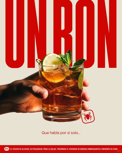 This is a vertically oriented promotional poster featuring a photorealistic, close-up image of a hand holding a short, clear glass filled with an amber-colored cocktail, garnished with a lime wheel and fresh mint leaves. The composition is centered on the glass, which is held from the left side by a person's hand with visible skin texture and warm, directional lighting. The background is a solid, warm off-white or beige, creating a clean, studio-like setting that isolates the subject. The dominant color palette is warm and inviting, centered on the rich amber and copper tones of the drink, accented by the vibrant green of the mint and lime, all set against the neutral backdrop. The lighting is soft but directional, coming from the upper left, creating gentle highlights on the glass, ice, and hand, and casting subtle shadows that give the scene depth and a tactile, three-dimensional feel. The texture is highly realistic, with the condensation on the glass, the facets of the ice cubes, the fibrous surface of the mint leaves, and the skin details all rendered with high fidelity. The composition uses a shallow depth of field, keeping the glass and hand in sharp focus while the background remains completely flat and unobtrusive. The shape language is a contrast between the strong, geometric lines of the large red text at the top and the organic, irregular forms of the hand, lime, and mint. The mood is sophisticated, refreshing, and slightly indulgent, conveying a sense of casual elegance. The text is a major visual element: large, bold, sans-serif letters in a saturated red spell "UNRON" at the top, dominating the upper third of the image. Below the glass, smaller red text in a clean sans-serif font reads "Que habla por sí solo..." (Spanish for "That speaks for itself..."). At the very bottom, a red banner contains white text in all caps: "EL EXCESO DE ALCOHOL ES PERJUDICIAL PARA LA SALUD. PROHÍBASE EL EXPENDIO DE BEBIDAS EMBRIAGANTES A MENORES DE EDAD." (Spanish for "Excessive alcohol is harmful to health. The sale of intoxicating beverages to minors is prohibited."). A small red square icon with a stylized spider logo is positioned to the lower right of the glass. The focus is squarely on the cocktail as the hero product, with the lighting and composition designed to make the drink appear appealing, cold, and meticulously prepared.