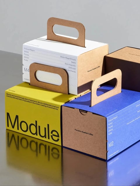 The image displays a studio-style product photograph featuring three distinct coffee packaging boxes, each designed as a portable carrier with integrated handles, arranged in a stacked and overlapping composition on a reflective dark gray surface against a neutral light gray background. The overall visual aesthetic is clean, minimalist, and photorealistic, emphasizing the tactile material quality of the packaging. The color language is defined by a high-contrast, primary-inspired palette: one box is a vibrant, saturated mustard yellow, another is a deep royal blue, and the third is a clean white with natural kraft brown cardboard handles and interior accents. The lighting approach utilizes soft, diffused directional light from the upper left, casting gentle shadows that define the forms and create a polished, commercial mood with subtle highlights on the glossy surfaces. Texture and material feel are central, with a clear distinction between the smooth, matte-finish printed cardstock of the outer panels and the rough, fibrous texture of the unbleached kraft cardboard handles and interior edges. Compositionally, the framing is a close-up, slightly high-angle shot that fills the frame, with the boxes creating a diagonal arrangement that guides the eye, and the reflective surface adds depth and symmetry. Shape language is predominantly geometric and architectural, featuring sharp right angles, clean lines, and the repetitive, functional oval cutouts of the handles. Visible text includes the large, bold sans-serif word "Module" on the yellow box, smaller descriptive text on the white and blue boxes listing coffee details like "Producer/Region," "Varietal," "Gesha Village/Ethiopia," "Process," and "Precision Roasted Coffee," all in a clean, modern sans-serif typeface in English, which serves a functional and branding role. The overall mood is modern, artisanal, and precise, conveying a sense of premium quality and careful design through its structured arrangement and clean material contrasts.