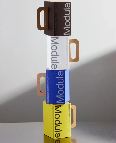 This image is a photorealistic 3D render showcasing a vertical stack of four rectangular cardboard boxes, each with a handle cutout and differing in color. The composition is centered against a plain, light gray gradient background, with a soft shadow cast to the left, indicating a light source from the upper right. The top box is dark brown, the second is white, the third is blue, and the bottom is yellow. Each box features the word "Module" printed vertically in large, clean, sans-serif typography; the text color contrasts with the box color (white text on brown and blue, gray on white, black on yellow). Alongside the large text, smaller descriptive paragraphs in English are printed vertically on the side panels of each box. The visual aesthetic is minimalist and modern, characterized by clean geometric shapes, sharp edges, and smooth, matte material surfaces. The lighting is soft and diffused, creating subtle gradients and a sense of depth without harsh highlights. The color palette is a striking combination of saturated primary and neutral tones (brown, white, blue, yellow) against a neutral background, creating a vibrant yet orderly mood. The arrangement emphasizes modularity and product design through its repetitive yet color-varied form.