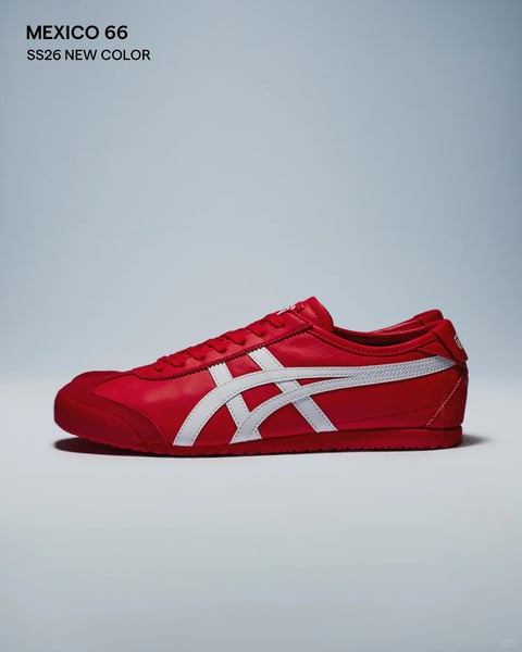The image features a single Onitsuka Tiger Mexico 66 sneaker as the central subject, presented in a clean, studio-style product shot. The composition is a side-profile view, centered horizontally against a smooth, neutral light grey gradient background that transitions subtly from a lighter tone at the top to a slightly darker, grounded grey at the bottom, creating a sense of depth and focus on the shoe. The sneaker itself is rendered in a vibrant, saturated crimson red leather, with a matching red suede toe cap and a red rubber sole. The iconic Asics stripes on the side are a crisp, contrasting white. In the upper left corner, there is small, clean, black sans-serif text arranged in two lines: the top line reads 'MEXICO 66' and the line below reads 'SS26 NEW COLOR'. This text is minimal, informative, and does not interfere with the visual presentation of the product. The lighting is soft and diffuse, appearing to come from the front and slightly above, casting a gentle, subtle shadow beneath the shoe that grounds it on the surface without creating harsh contrasts. The overall visual aesthetic is photorealistic and polished, emphasizing the shoe's design details, materials, and color through a minimalist, high-contrast approach. The dominant color language is a bold, high-saturation red paired with stark white and neutral grey, creating a vibrant, energetic, and modern mood with a strong graphic quality.