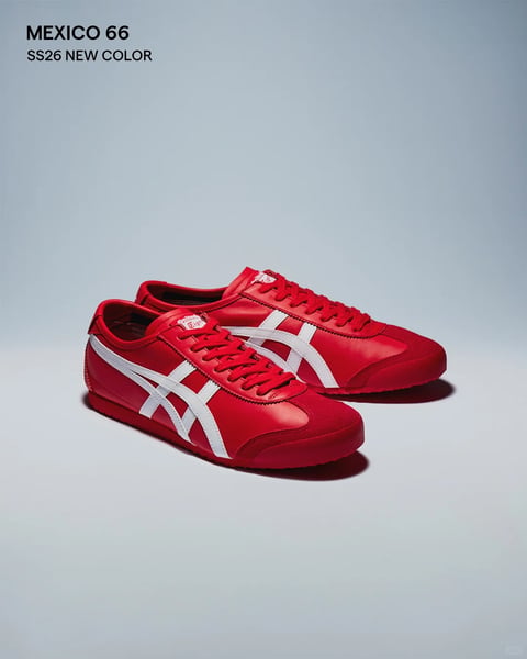 The image presents a photorealistic product shot of a pair of Onitsuka Tiger Mexico 66 sneakers, positioned as the central subject against a plain, gradient background that transitions from a light, cool blue-grey at the top to a slightly warmer, lighter grey at the bottom. The shoes are arranged in a dynamic, slightly staggered composition, with the left shoe placed forward and angled towards the viewer, and the right shoe behind it, creating a sense of depth and showcasing the side profile. The dominant color palette is a vibrant, saturated red for the shoe's leather upper, laces, and sole, sharply contrasted by crisp white for the iconic Asics stripe detailing and tongue label. Text appears in the top left corner in a clean, sans-serif font, reading 'MEXICO 66' in bold black and 'SS26 NEW COLOR' in a smaller, regular weight, indicating the model and a seasonal color release. The lighting is soft and directional, coming from the upper left, casting gentle, diffused shadows to the lower right of the shoes, which highlights the smooth texture of the leather and the subtle contours of the suede toe cap. The materials are rendered with a realistic tactile quality, emphasizing the sleek leather, the finer grain of the suede, and the matte rubber of the sole. The overall mood is clean, modern, and focused, with a high-contrast, studio-quality aesthetic designed to highlight the product's design and new colorway.