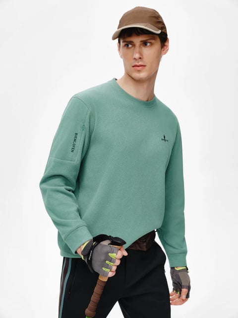 The image presents a photorealistic studio shot of a young male model positioned against a plain, bright white background, with a medium framing that captures him from the mid-thigh up as he looks off-camera to the left. He is styled in a relaxed, sporty outdoor aesthetic, wearing a muted sage-green crewneck sweatshirt, black athletic pants with vertical stripe detailing in brown and teal, and fingerless gray and neon yellow gloves, while holding the cork-grip handle of a trekking pole in his right hand. Visible text appears in two locations on the sweatshirt: vertically oriented black text reading 'BIEMLOFEN' on the upper left sleeve, and a small black logo with text (appearing as 's@t&er' or similar) on the left chest, both serving as subtle brand identifiers. The lighting is soft and even, typical of commercial photography, creating gentle shadows that define the model's facial features and the soft, matte texture of the fleece sweatshirt without harsh contrast. The color palette is dominated by the cool, desaturated green of the top, anchored by the neutral black pants and accented by the warm brown of the cap and trekking pole grip, alongside the vibrant pop of neon yellow on the gloves, resulting in a natural, earthy yet modern color scheme. The composition is clean and centered, emphasizing the clothing and accessories with clear spatial arrangement, while the textures are rendered with high detail, from the plush, soft-touch surface of the sweatshirt to the ribbed knit of the gloves and the smooth, technical fabric of the pants. The overall mood is calm and focused, conveying a sense of prepared, understated outdoor activity with a contemporary athletic edge.